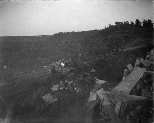 Elevated view over wooden scaffolding towards several plows on a road pulled by horses. In the background are tree-covered hills.
