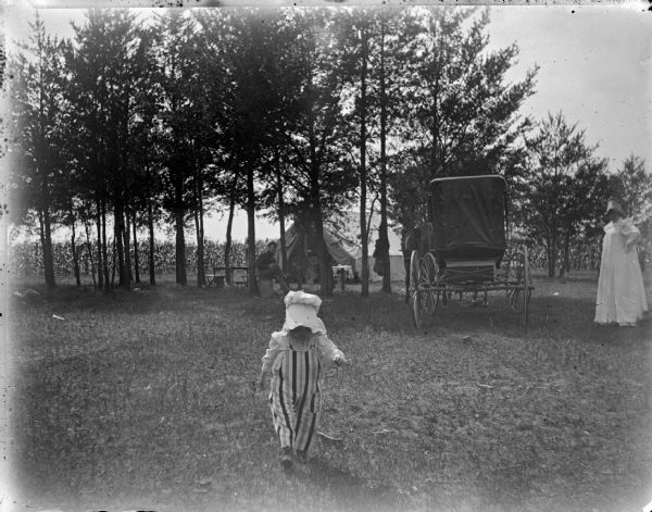 View towards a small child walking in the foreground, wearing a bonnet and striped overalls. In the background on the right near a wagon is a woman in a white dress and bonnet standing and watching the child. In the background under trees is a man sitting. Behind the trees is a a tent.