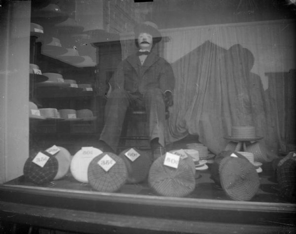Outdoor view of a store window showing a mannequin and hats for sale.