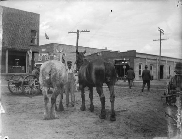 Outdoor view of a man posing standing and displaying two horses on a city street, with several buildings and men in the background.