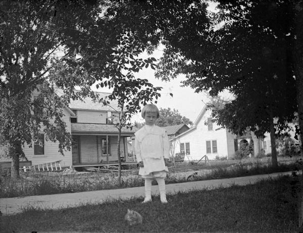 Outdoor view of a small child posing standing near a wooden walkway. Houses are across the street in the background. A kitten is resting in the grass in the foreground.