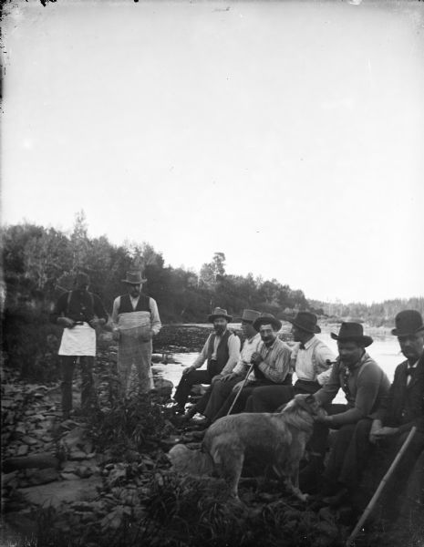 Outdoor view of a group of men posing standing and sitting near the shore of a river with a dog.