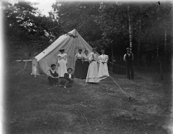 Outdoor view of five women and a man posing standing, and a woman and a man on the ground near a tent with the acronym "B.R.F.D.A." printed on it.