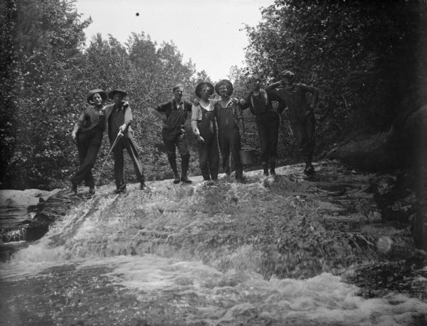 Group portrait looking up towards a group of young men posing standing  on a large rock waterfall of a river. One man is holding a fishing pole and another man has a pail. Many of the men have pipes in their mouths.
