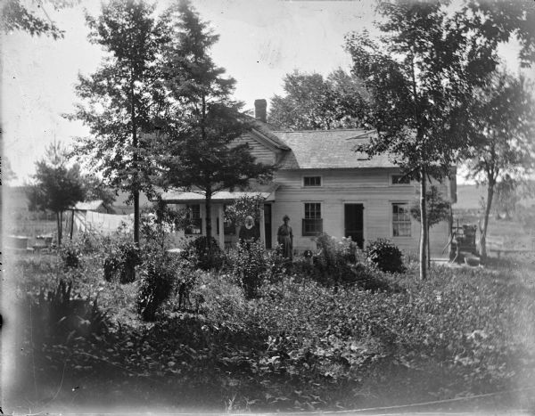 View towards two women and a dog posing standing in the yard of a wooden house. Trees and plants fill the yard, and on the far left laundry is hanging from a clothesline. On the far right is a hand pump.