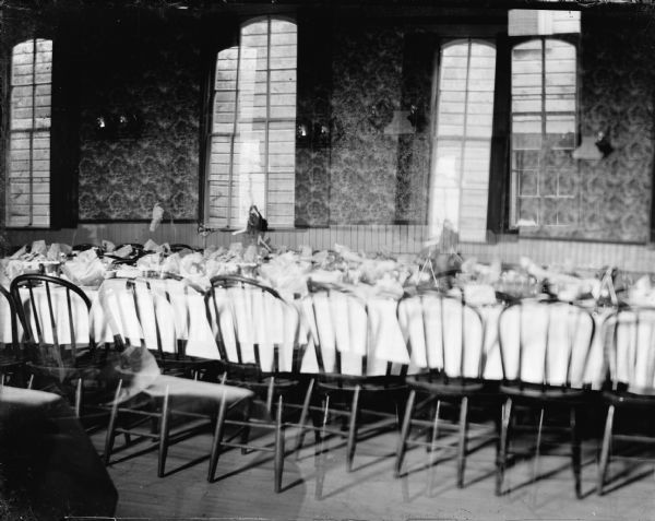 Long tables are set for a meal in a dining hall. Tall windows are in the wall in the background, with a view of an exterior wall of another building outdoors. The double exposure is of the same view of tables and windows.