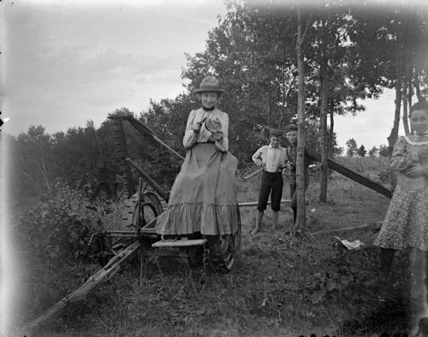 Outdoor view of a woman posing sitting on a mower in front of two boys posing standing, and another woman posing standing on the right.