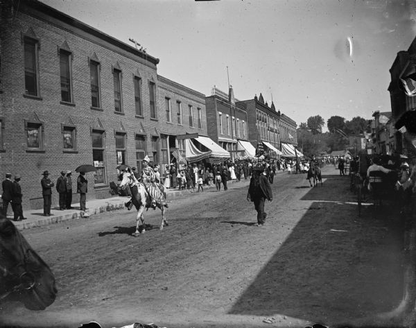 Outdoor view of a girl in a patriotic costume riding a horse on a town street lined with people. Further down the street are other individuals in the parade. Identified as a Memorial Day parade in Black River Falls.