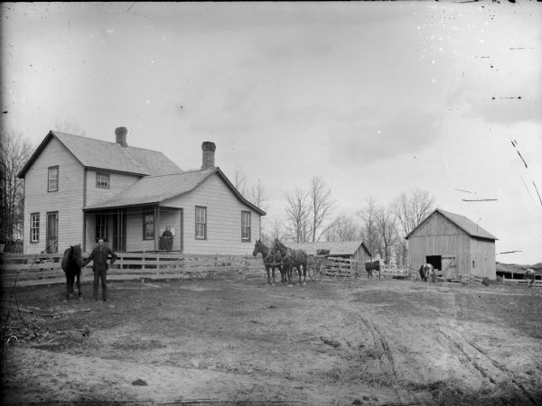 View of a farm, with a man posing with a horse on the left in front of a fence. A woman is sitting on the porch of the farmhouse. Two horses with a buggy are in the center. A group of cows are standing on the right by the stable.