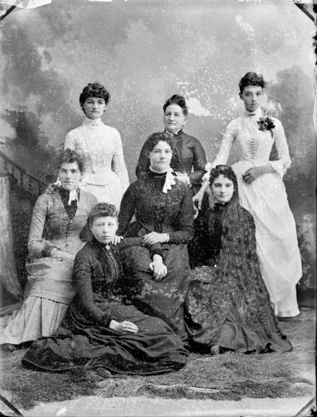 Studio group portrait in front of a painted backdrop of seven unidentified European-American women. The four women in front are sitting; the three women in the back are standing.