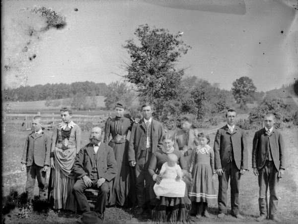 Outdoor group portrait of men, women and children, perhaps an extended family. The oldest couple, a man and woman, is seated in the center, with the woman holding an infant in her lap.