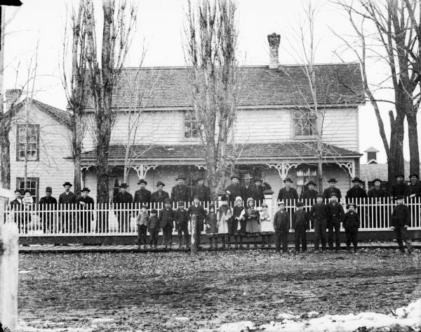Outdoor group portrait of a group of men and children. The children, boys and girls, are standing on a wooden sidewalk in front of a picket fence, and a line of men are standing behind the fence. Behind them is a house. A man on the far left is holding an infant, and some of the girls are holding dolls.