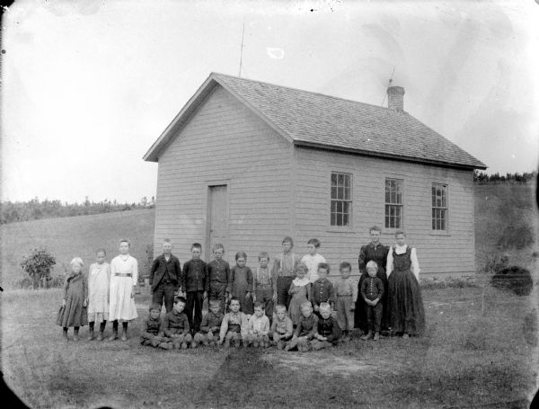 Outdoor group portrait of students and teachers posing for a class portrait in front of a one-room schoolhouse.