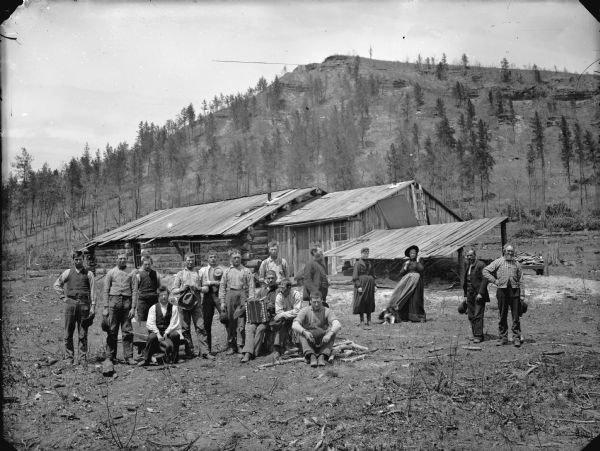 Outdoor group portrait of a people gathered in front of a log cabin. A dog is next to one of the women in the back. The man in the center has an accordion on his lap. A steep hill with pine trees is in the background.