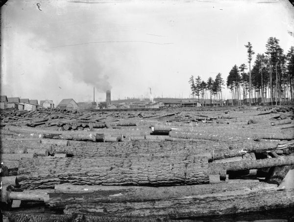 View of logs stacked closely together. Industrial buildings and trees are in the background.