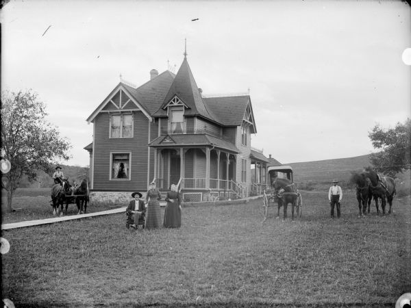 A group of people and horses are assembled on the lawn of a multi-gabled house with a wooden sidewalk. There are a few trees in the yard, and a slope in the background.