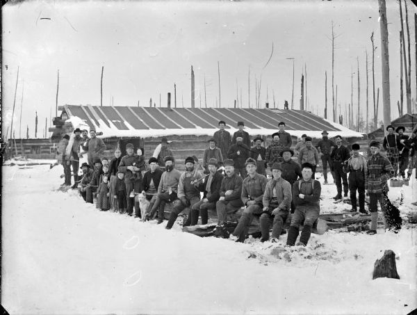 Outdoor group portrait of men posing together at a logging camp. There is also a family with five children, and a dog in the center.