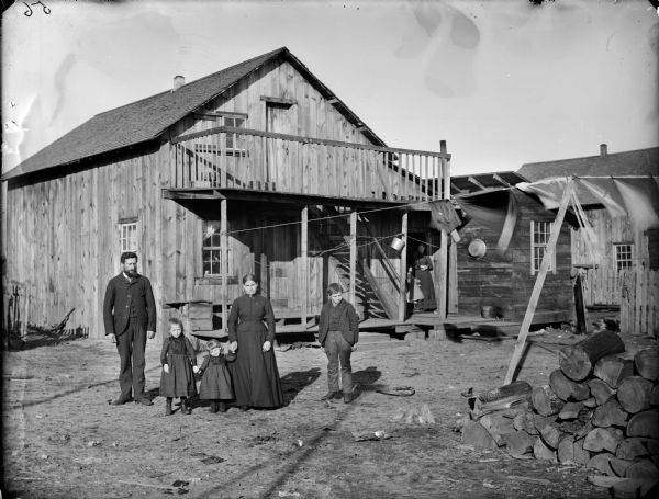 Outdoor group portrait of a man, two girls, a woman and a boy standing in the yard in front of a two-story wooden house. Another woman and girl are standing at an open doorway of the house under a balcony on the right. Laundry is handing from a clothesline across the yard, and fuelwood is stacked in the right foreground.
