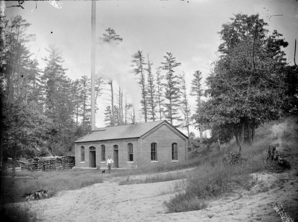 View across open area towards a brick power plant, where a man and a small dog are standing. Cords of wood are stacked at the left side of the building. On the far right is a steep, dirt road lined with trees.