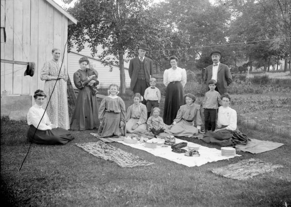 Outdoor group portrait of a group having a picnic in a garden. Women and children are sitting around blankets on the ground, and women and men are standing behind them. There are wooden buildings behind the group on the left, and horses are in the far background under trees.
