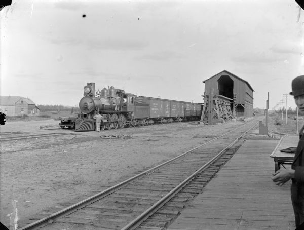 View from a railroad platform and railroad tracks. A locomotive with railroad cars is parked on the far track. One man is standing next to the locomotive; another man is standing on the platform in the right foreground.