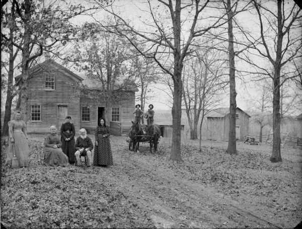 Outdoor group portrait of four women and three men posing in front of their farmhouse. Two of the men are standing on a cart pulled by two horses. The trees are bare of leaves.