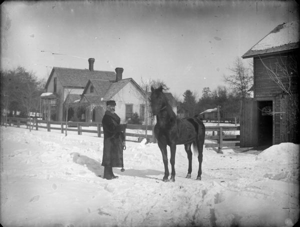 Man in a fur coat displaying a horse on a road covered in snow. There is a barn on the far right, and a house in the background.
