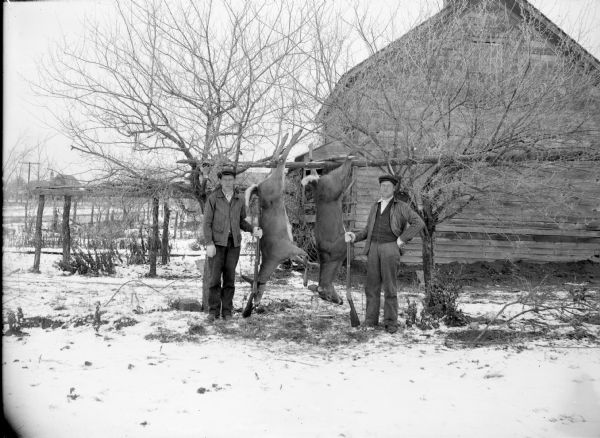 Outdoor view towards two deer hunters posing with their rifles and two deer carcasses. Snow is on the ground, and in the background is a wooden building.