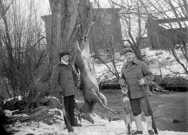 Outdoor portrait of two hunters posing with a deer carcass, a fox carcass and rifles on the edge of a stream in winter with snow on the ground. Building are across the river in the background.