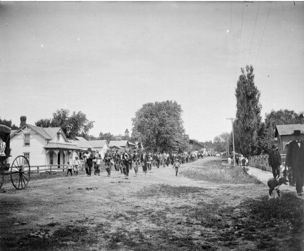 View down unpaved road towards a band at the head of a parade coming down a town street.