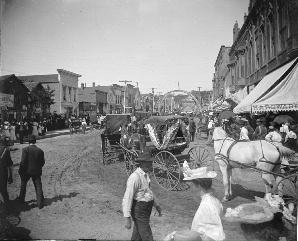 Slightly elevated view of a parade on a town street. Identified as a parade in Black River Falls, taking place for the Wisconsin State Fair.