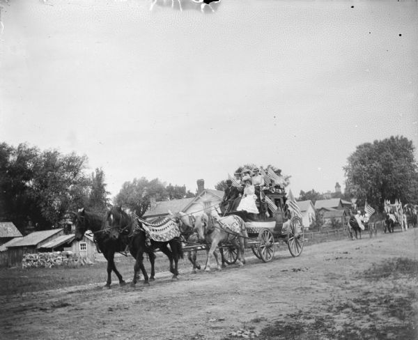 View across unpaved road towards a group of people on a stagecoach patriotically decorated for a parade on a town street.