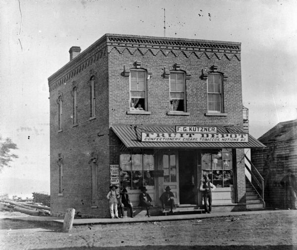 Copy photograph of a storefront. The store is identified as F.G. Kutzner Fruit Depot. Men are standing and sitting out front. Children are looking out of the second story window.