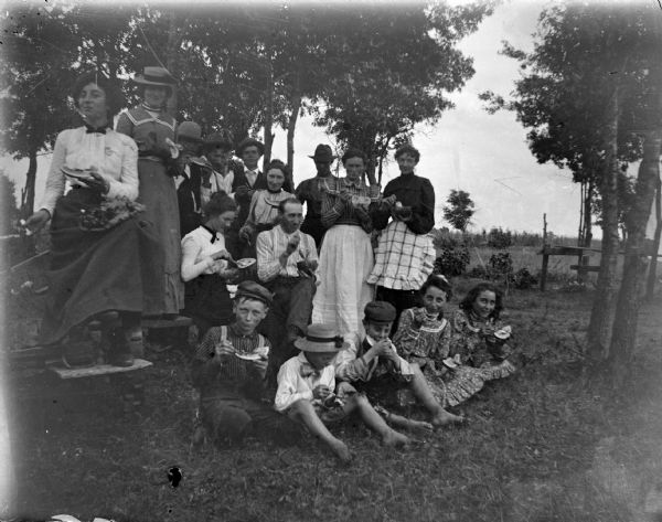 Outdoor portrait of a large group of men, women, and children posing sitting and standing and eating watermelon.