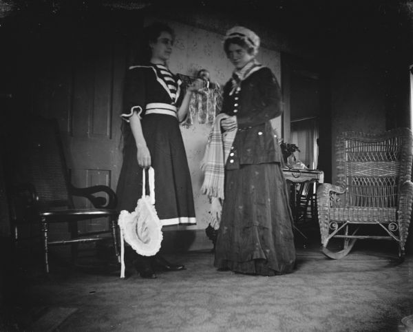 Interior portrait of two women posing standing indoors near two chairs. The woman on the left is holding an apple and a bonnet, and the woman on the right is holding a plaid scarf.