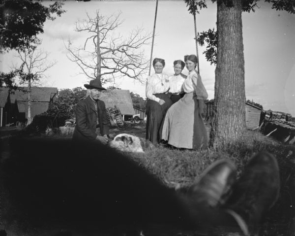 Outdoor group portrait of three women posing sitting on a swing under a tree. There is a man posing sitting with a dog in front of them. In the background are farm buildings. In the foreground are the legs and shoes of the photographer.