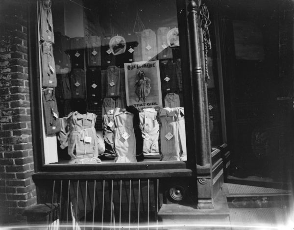 View from sidewalk of a storefront window displaying shirts. The display includes a poster for "Olga Lorraine in 'That Girl' at the Opera House."
