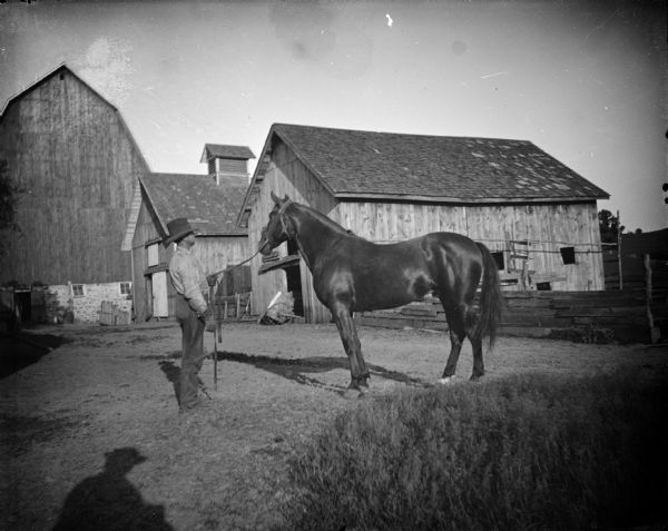 Outdoor portrait of a man posing standing and displaying a horse in front of wooden buildings and a barn.