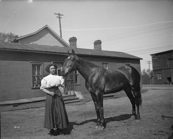 Outdoor portrait of a woman posing standing and displaying a single horse on a street in front of a wooden buildings.
