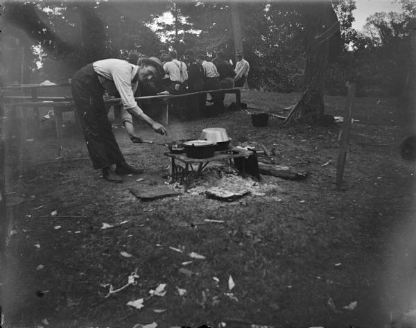 Outdoor view towards a man leaning over a campfire, with a group of men sitting at a picnic table in the background.
