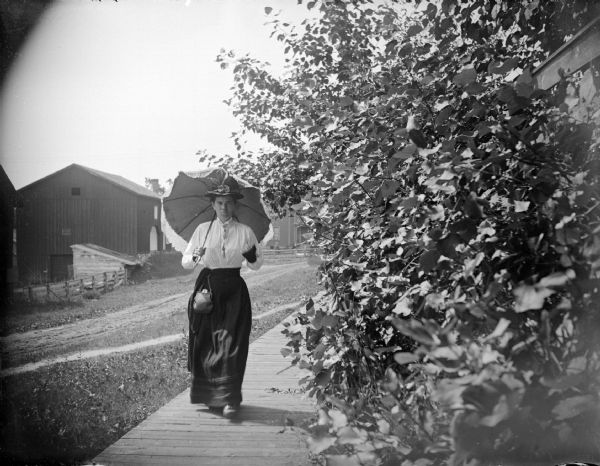 View down wooden sidewalk towards a woman walking and holding a parasol. There are wooden buildings in the background.