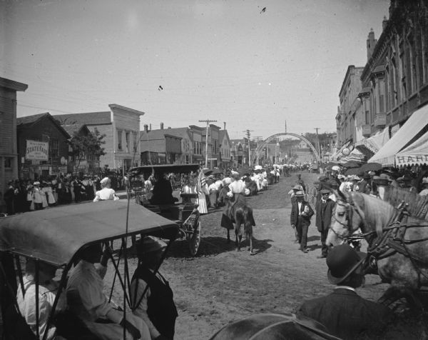 Slightly elevated view of a parade on a city street. Location identified as Main Street in Black River Falls, for the State Fair in 1905.