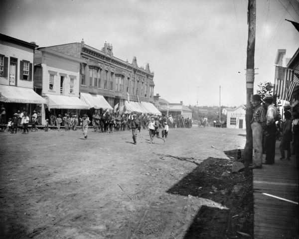 View down unpaved street towards children walking along near a band marching in front of a group of people in a parade. People are watching from a shaded sidewalk on the far right
