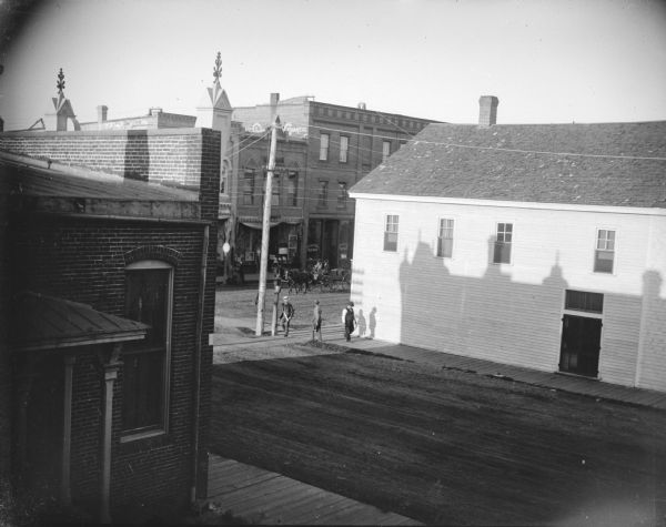 Elevated view of an intersection of two streets. Men are walking on the wooden sidewalks, and a wagon is moving down the street. Location identified as the intersection of Main and First Streets from the window of the photographic studio of Charles J. Van Schaick.