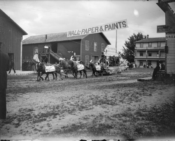 View across unpaved street towards a wagon pulled by a team of six horses advertising "The People's Drugstore" under a banner that reads "Wall Paper and Paints." The horses have banners that read: "Use Gold Band Flour." Location identified as the street in front of the Merchants Hotel in Black River Falls.