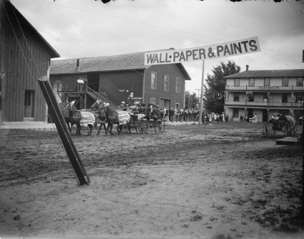 Exterior portrait of a wagon pulled by a team of four horses and under a banner that reads "Wall Paper and Paints." A marching band is gathered on a sidewalk. Location identified as the street in front of the Merchants Hotel in Black River Falls.