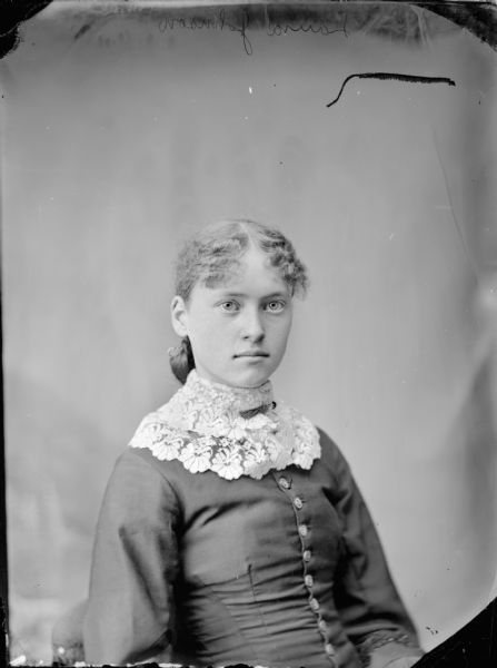 Waist-up studio portrait of a young woman posing sitting and wearing a dark-colored button-up blouse with a wide light-colored lace collar. Identified as probably Laura Johnson.