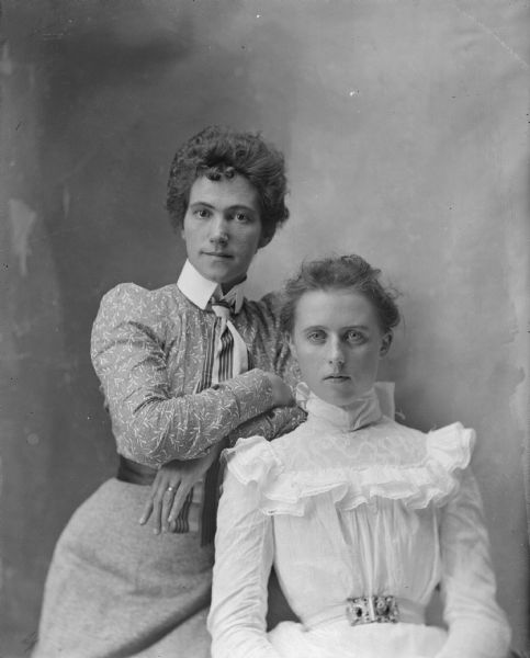 Studio portrait of two women. The woman on the left is standing and leaning to the right and is wearing a dark-colored blouse and skirt. The woman on the right is sitting and is wearing a light-colored dress.