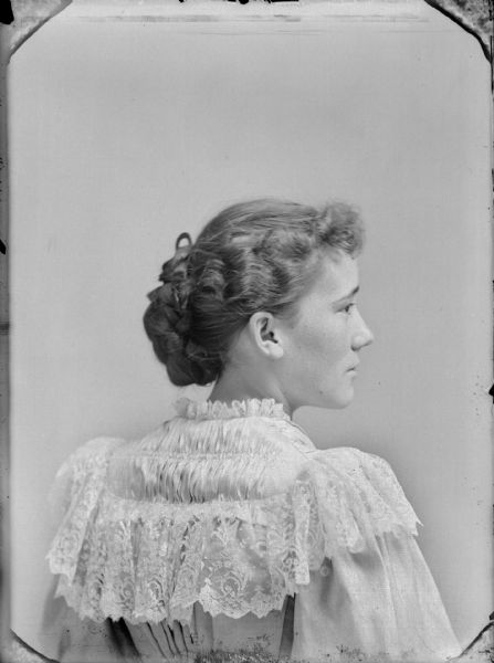 Over the shoulder studio portrait of a woman looking to the right in profile. She is wearing a light-colored blouse with lace trim.