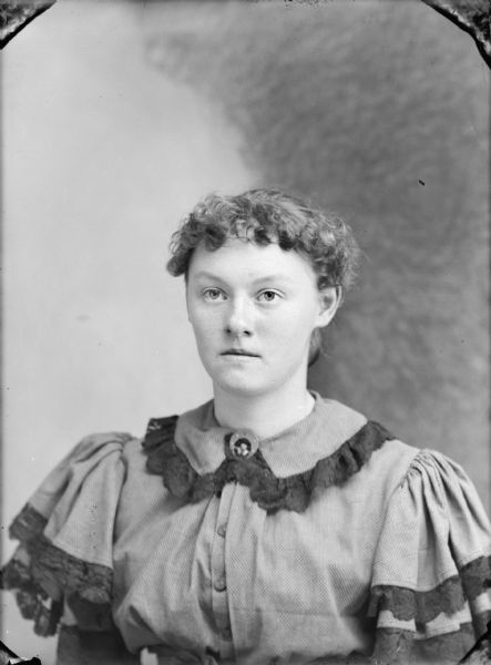 Waist-up studio portrait of a woman. She is wearing a light-colored dress with dark-colored lace trim, and a collar pin.